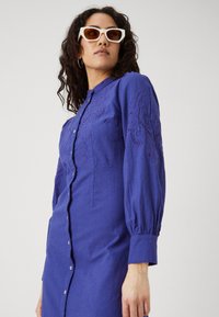 Vestito in lino blu con dettagli ricamati, chiusura a bottoni e maniche a sbuffo. Abbinato a occhiali da sole bianchi angolari. Tessuto testurizzato.