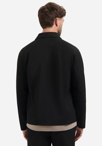 Mann mit kurzen braunen Haaren, der eine schwarze Jacke über einem beigen Hemd trägt, von hinten gegen einen schlichten hellen Hintergrund gezeigt.