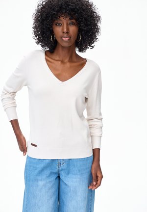 Kobieta o kręconych włosach, nosząca złote kolczyki koła, biały sweter z dekoltem w serek i jasnoniebieskie dżinsy, na tle jednolitej białej ściany.