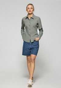 Ljusgrön knäppt skjorta med långa ärmar, kombinerad med mörka jeansshorts. Modellen bär off-white sneakers. Bakgrunden är neutral grå.