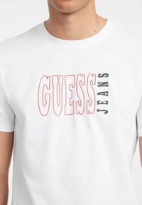 Balta medvilninė marškinėliai su ryškiai raudonu ir juodu grafiniu užrašu "GUESS JEANS". Trumpomis rankovėmis, apvaliu iškirpimu, lygiu paviršiumi.
