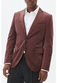 Burgunderfarbener Blazer aus weichem Stoff, mit Reverskragen, zwei Knöpfen, seitlichen Taschen und einer einzelnen Brusttasche. Darunter ein weißes Hemd.