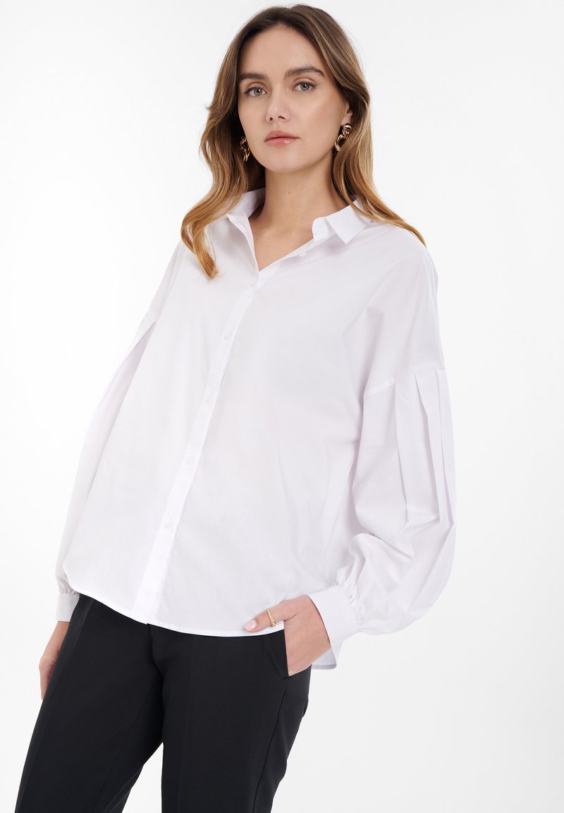 Bílá košile s button-up designem, uvolněného střihu, s dlouhými nabíranými rukávy a hladkou texturou; doplněná černými zúženými kalhotami.