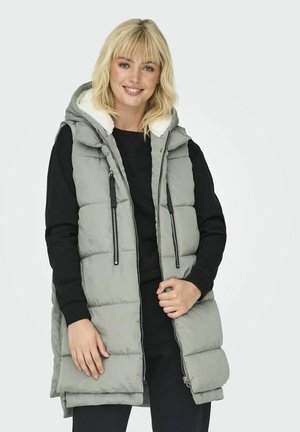 Doudoune grise sans manches avec capuche et doublure en polaire, dotée de deux fermetures éclair latérales et de cordons de serrage ajustables. Portée par-dessus un pull noir.