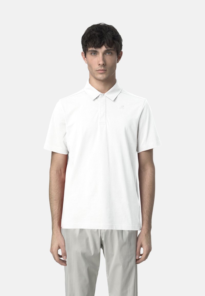 Polo bianco con colletto e maniche corte, caratterizzato da un leggero dettaglio con logo, abbinato a pantaloni grigio chiaro.
