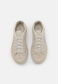 Béžové plátěné tenisky s texturovaným povrchem, bílé tkaničky a fialové detaily, s plochou gumovou podrážkou a kulatým tvarem špičky.