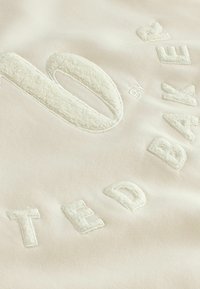 Beige stof met verhoogde, gestructureerde letters die "TED BAKER" spellen en een grote, gestructureerde nul in het midden.