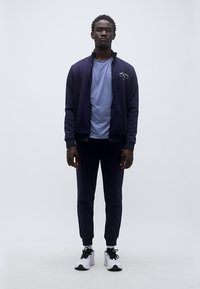 Ναυτικό φόρμα με ζακέτα με φερμουάρ, μακριά μανίκια και ταιριαστό παντελόνι. Φοράει γαλάζιο μπλουζάκι από μέσα. Το μοντέλο φοράει λευκά αθλητικά παπούτσια.