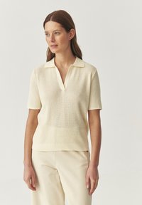 Maglietta a maniche corte in stile polo di un beige chiaro, tessitura in maglia, con scollo a V e colletto a contrasto. Abbinata a pantaloni beige chiaro.