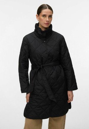 Mujer con un abrigo acolchado negro de cuello alto y cinturón atado, de pie contra un fondo claro y sencillo.