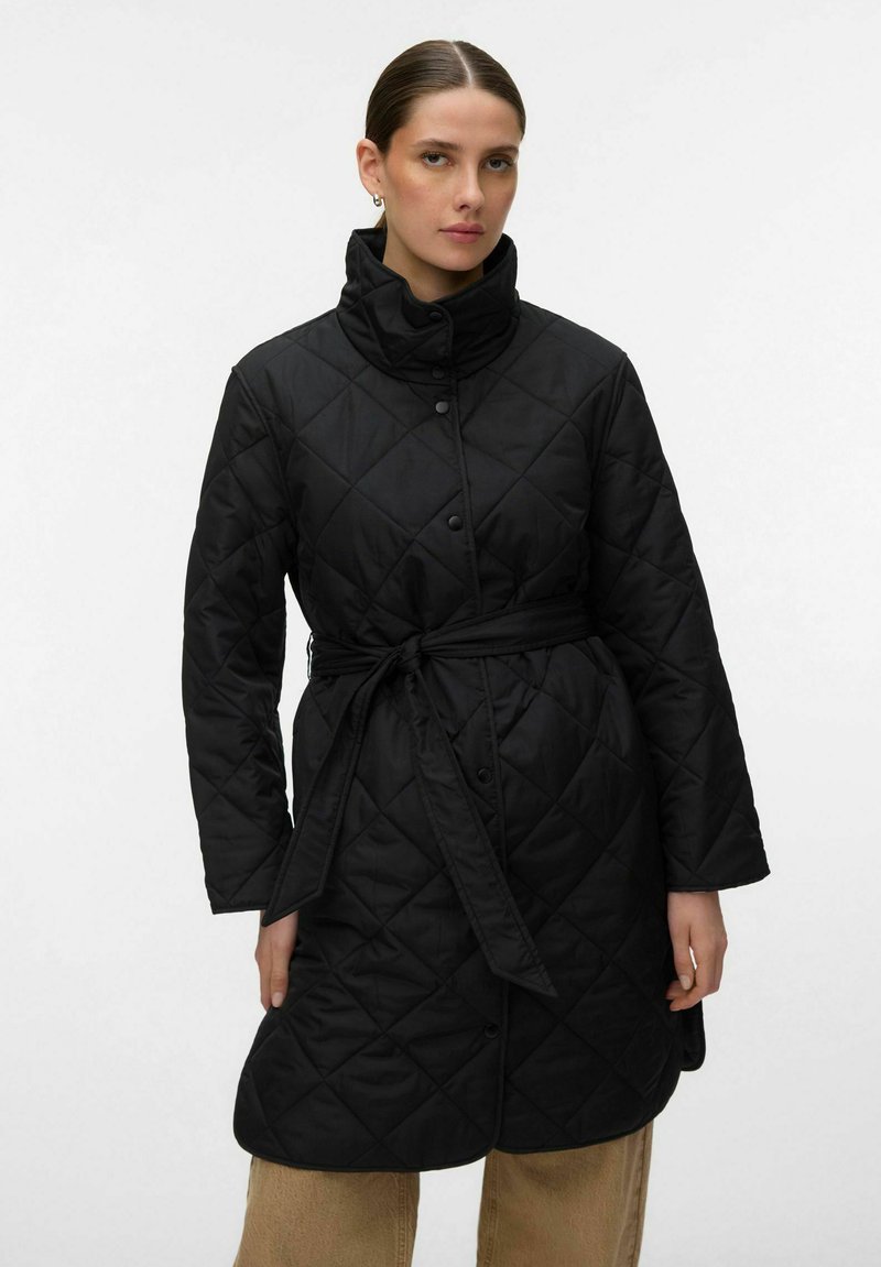 Ženska, oblečena v črno prešito plašč z visokim ovratnikom in pasom, stoji pred enostavnim svetlim ozadjem.