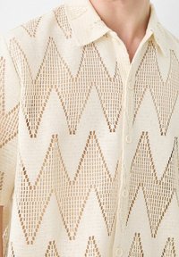 Maglietta in maglia a trame aperte color crema con motivo a zigzag. Maniche corte, colletto e chiusura frontale con bottoni. Tessuto strutturato con un design leggero e traspirante.