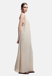 Beige maxiklänning med en krinklad textur, med ärmlös design och avslappnad passform. Modellen står i profil och visar upp den flödande siluetten.