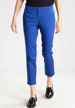 Vrouw draagt felblauwe, nauwsluitende, cropped broek en zwarte loafers met franjes, staand tegen een effen witte achtergrond.