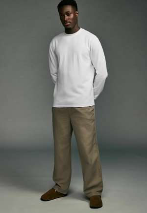 Hvid langærmet skjorte, khaki bukser og mørkebrune slippers. Modellen står med hænderne bag ryggen, mod en neutral grå baggrund.
