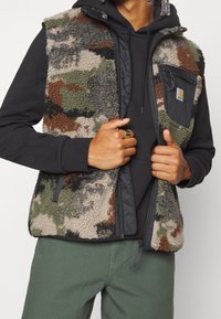 Chaleco de forro polar con un patrón de camuflaje en verde, marrón y gris. Presenta un bolsillo frontal con cremallera negra y un cuello alto.