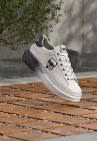 Λευκά δερμάτινα αθλητικά παπούτσια με μαύρη λεπτομέρεια και λογότυπο καρτούν. Διαθέτουν στρογγυλή μύτη, δέσιμο με κορδόνια μπροστά και υφασμένο, βαθμιαίο πέλμα.