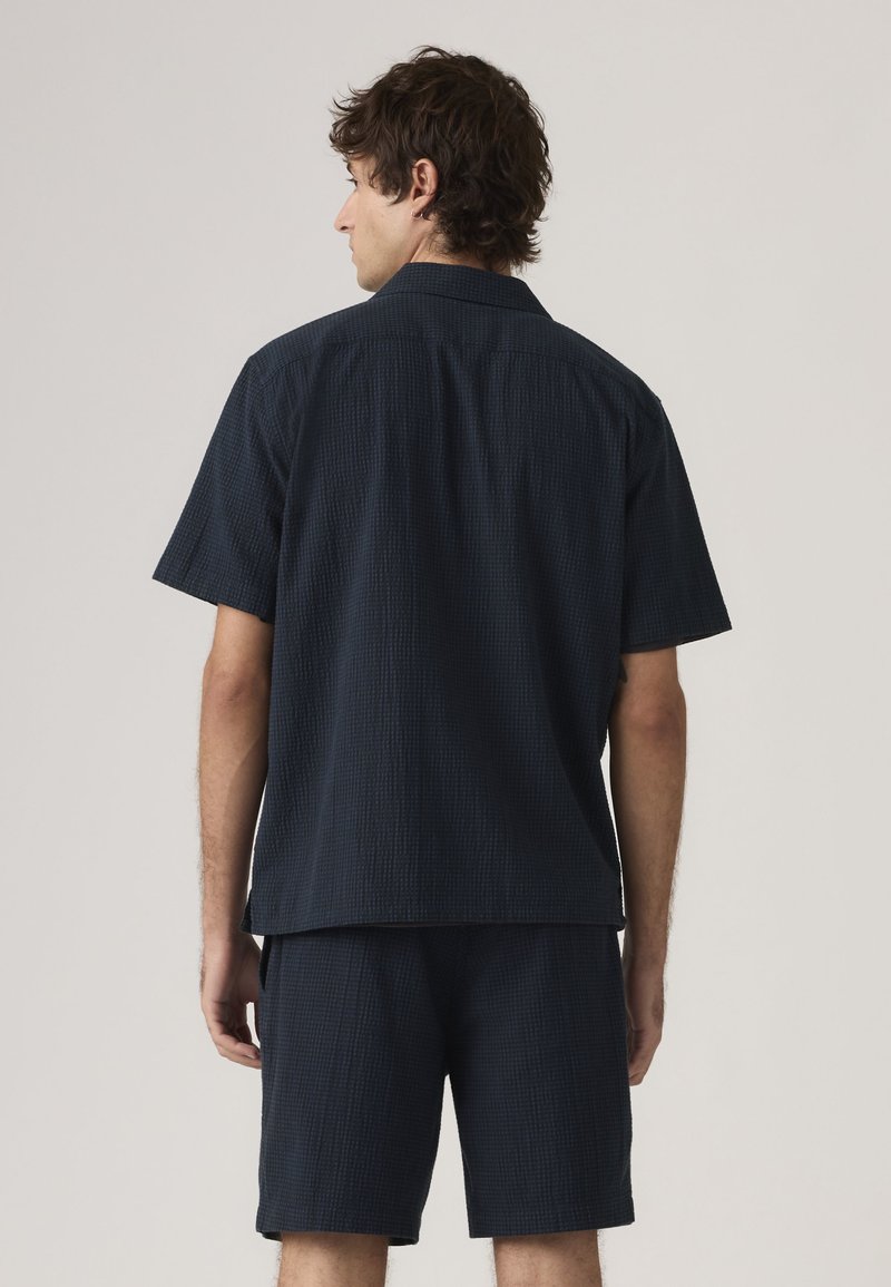 Mand med mørkt krøllet hår iført matchende løstsiddende mørkeblå kortærmet skjorte og shorts, set bagfra mod en ensfarvet lys baggrund.