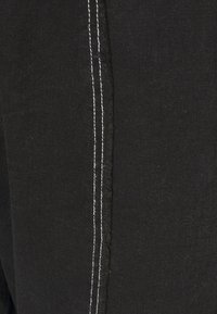 Tecido de denim preto com costura branca ao longo da costura lateral; apresenta bordas desfiadas. Perspectiva de close-up destacando a textura e os detalhes.