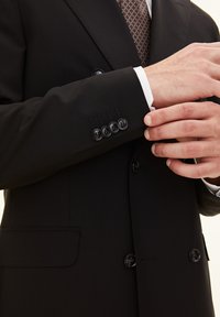 Zwarte op maat gemaakte colbert met een dubbele knoopsluiting, vier zwarte knopen op de manchet en een wit overhemd dat onder de mouw vandaan komt.