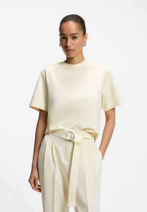Femme aux cheveux courts tirés en arrière, portant un haut crème à manches courtes et un pantalon crème taille haute avec une ceinture épaisse, posant devant un fond uni.