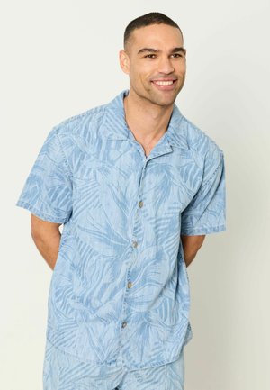 Lächelnder Mann mit hellblauem Kurzarmhemd mit dezentem Blattmuster, stehend mit den Händen hinter dem Rücken.