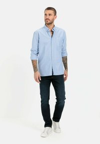 Lichtblauw overhemd met knopen, lange mouwen, enkele borstzak; gecombineerd met donkere jeans en witte sneakers, op een neutrale achtergrond.