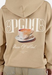 Sweatshirt bej cu un grafic mare al unei cești de cafea, cu textul „PGDF” și „Peace Of Mind” în alb contrastant. Croială moale, lejeră.