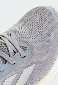 Chaussure de sport grise avec un upper en tricot texturé, des œillets renforcés et une semelle en caoutchouc blanc avec des accents gris angulaires.