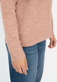 Rosa gestrickter Pullover mit geripptem Saum und Bündchen, der eine weiche, flauschige Textur aufweist. Kombiniert mit blauen Jeans, die eine Nahaufnahme des Ärmels zeigen.