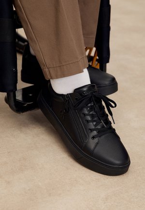 Baskets en cuir noir avec lacets et fermeture éclair latérale, portées avec des chaussettes blanches et un pantalon marron, le pied reposant sur le repose-pieds d'un fauteuil roulant.