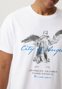 Biała bawełniana koszulka z grafiką skrzydlatej statuy, z napisem "Miasto Aniołów" w niebieskiej czcionce oraz "Los Angeles - Kalifornia" poniżej.