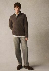 Brauner Wollpullover mit Kragen, über einem weiß gestreiften Hemd getragen. Hellgraue Jeans und braune Wildlederschuhe runden das Outfit ab.