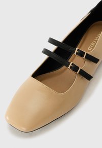 Beige nahkastossut, joissa on neliömäinen kärki, kahdella mustalla hihnalla, joissa on kultaiset soljet päällä, ja sileä pinta.