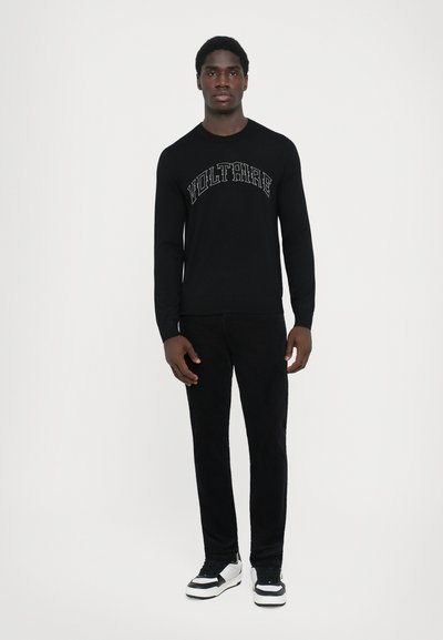 Sort sweatshirt med lange ærmer og teksten "VOLTAIRE", parret med sorte bukser og sneakers med hvide accenter, sat mod en enkel baggrund.