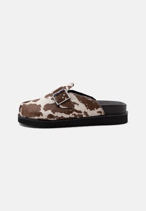 Παπούτσι τύπου slip-on με καφέ και λευκό σχέδιο μοτίβου αγελάδας, ασημί λουράκι με αγκράφα και μαύρη σόλα πάνω σε απλό φωτεινό φόντο.
