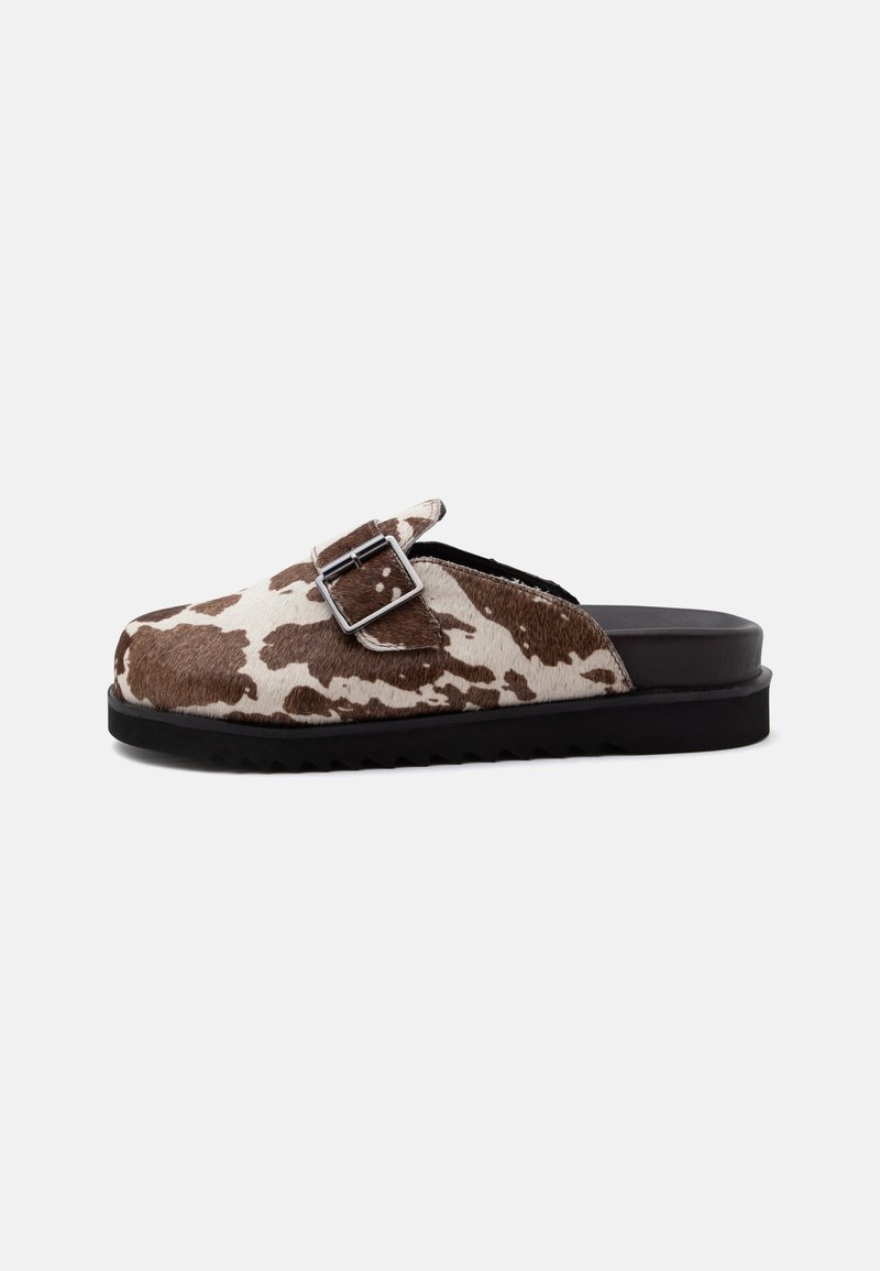 Chaussure slip-on avec motif vache marron et blanc, bride à boucle argentée et semelle noire sur fond clair uni.