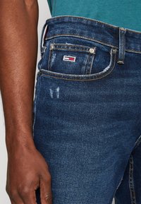 Niebieskie dżinsy o luźnym kroju, z widocznym przeszyciem, małą naszywką z logo na tylnej kieszeni oraz delikatnymi przetarciami.