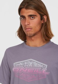 Grå kortærmet t-shirt med et stort pink og hvidt grafisk logo med indviklede mønstre. Stofniveauet ser blødt ud med en afslappet pasform.
