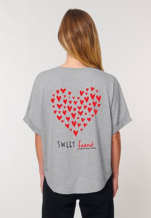 Une femme aux longs cheveux porte un T-shirt gris avec un grand cœur formé de cœurs rouges et le texte "SWEET heart" en noir et rouge.