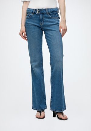 Γυναίκα που φοράει μεσαίου μπλε bootcut τζιν με διπλό κουμπί στη μέση και μαύρα σανδάλια με τακούνι, στέκεται μπροστά σε απλό ανοιχτό φόντο.