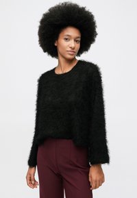 Afro frizurás nő egy fekete bolyhos, hosszú ujjú pulóverben és magas derekú bordó nadrágban áll, háttérben egy sima fehér fal.