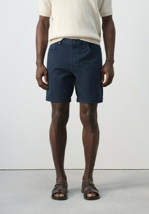 Hombre de piel oscura que lleva pantalones cortos azul marino con dobladillo enrollado, camiseta de punto de manga corta en color crema y sandalias de cuero marrón oscuro, de pie frente a un fondo liso.