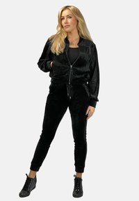 Czarny dres z weluru z kurtką na zamek i zwężającymi się spodniami. Teksturalny materiał o błyszczącym wykończeniu; połączony z czarnymi butami sportowymi na sznurówki.