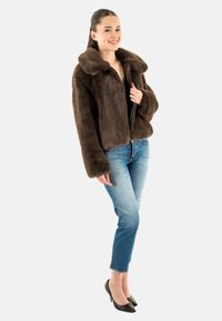Giacca in pelliccia sintetica marrone con colletto ampio, chiusura con zip e design accorciato. Abbinata a jeans skinny blu chiaro e tacchi neri.