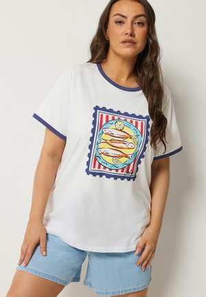 Kobieta z długimi brązowymi włosami, ubrana w białą koszulkę z niebieską lamówką i grafiką trzech ryb z plasterkami cytryny na talerzu, w połączeniu z jasnoniebieskimi szortami dżinsowymi.