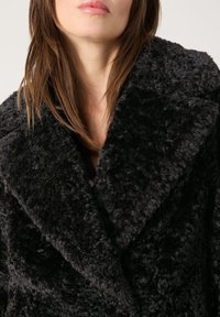 Abrigo negro de lana, doble botonadura, con una tela suave y texturizada; grandes solapas y un ajuste holgado, mostrando un vellón elevado y un color profundo.