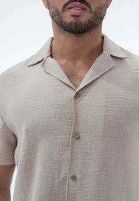 Chemise à manches courtes beige à boutons, fabriquée dans un tissu texturé, avec un col et deux boutons marron à l'avant.