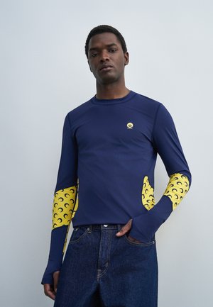 Bărbat purtând un tricou cu mâneci lungi de culoare albastru închis cu modele galbene în formă de semilună pe mâneci și buzunare, asortat cu blugi albastru închis, stând în picioare în fața unui perete simplu.