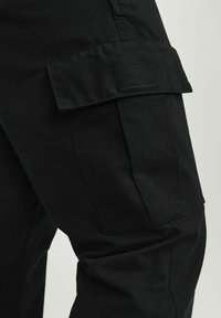 Czarne spodnie cargo wykonane są z wytrzymałej tkaniny, posiadają boczne kieszenie oraz prosty krój nogawek. Materiał ma gładką fakturę o matowym wykończeniu.